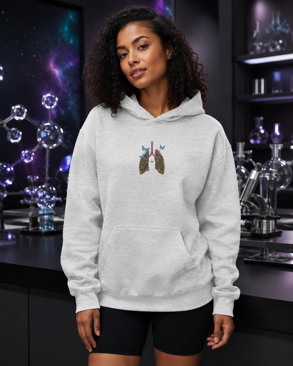 Young woman modeling the ash grey Butterfly Lungs hoodie in a premium science studio with glass molecular sculptures and cosmic lighting.