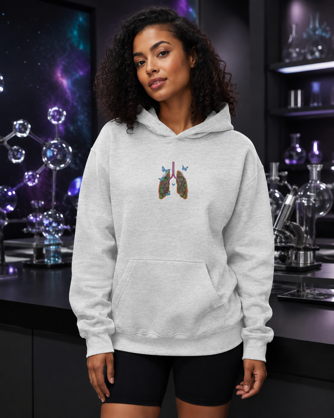Young woman modeling the ash grey Butterfly Lungs hoodie in a premium science studio with glass molecular sculptures and cosmic lighting.