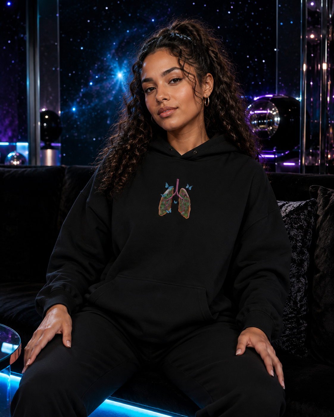 Young woman seated in a black Butterfly Lungs hoodie and black pants inside a luxurious violet-and-cyan galaxy lounge.
