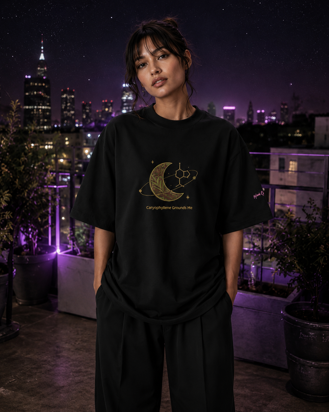 Woman wearing the black Caryophyllene Grounds Me oversized tee in a purple-lit rooftop garden overlooking the city.