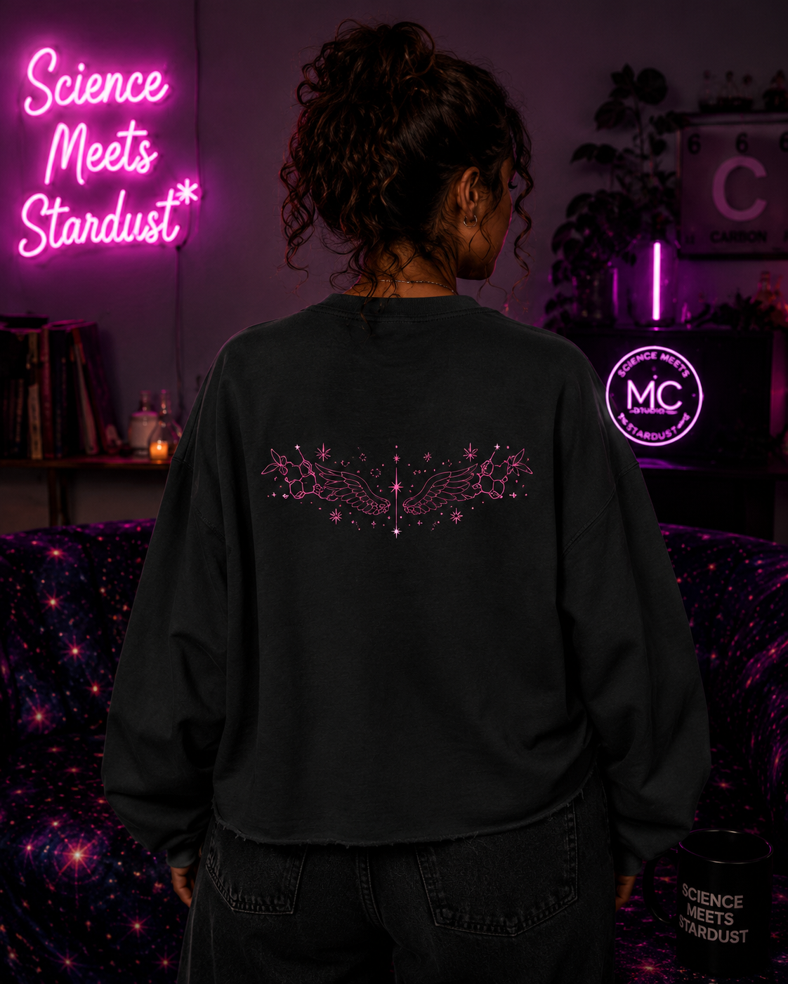 Back view of a woman wearing the Black Mandi Cruz Media raw edge boxy sweatshirt with pink celestial wing artwork in a Science Meets Stardust™ cosmic studio setting.