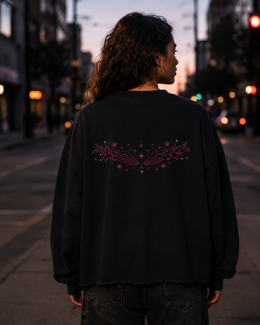 Back view of a woman wearing the Black Mandi Cruz Media raw edge boxy sweatshirt with pink celestial wing artwork on a city street at dusk.