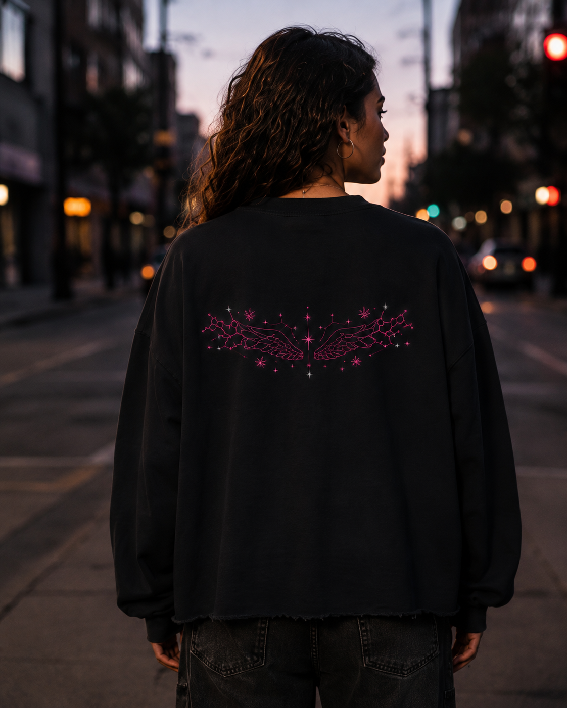 Back view of a woman wearing the Black Mandi Cruz Media raw edge boxy sweatshirt with pink celestial wing artwork on a city street at dusk.