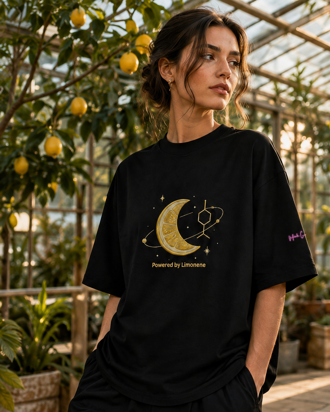 Woman wearing the black Powered by Limonene oversized high-neck tee in a sunlit conservatory filled with lemon trees.