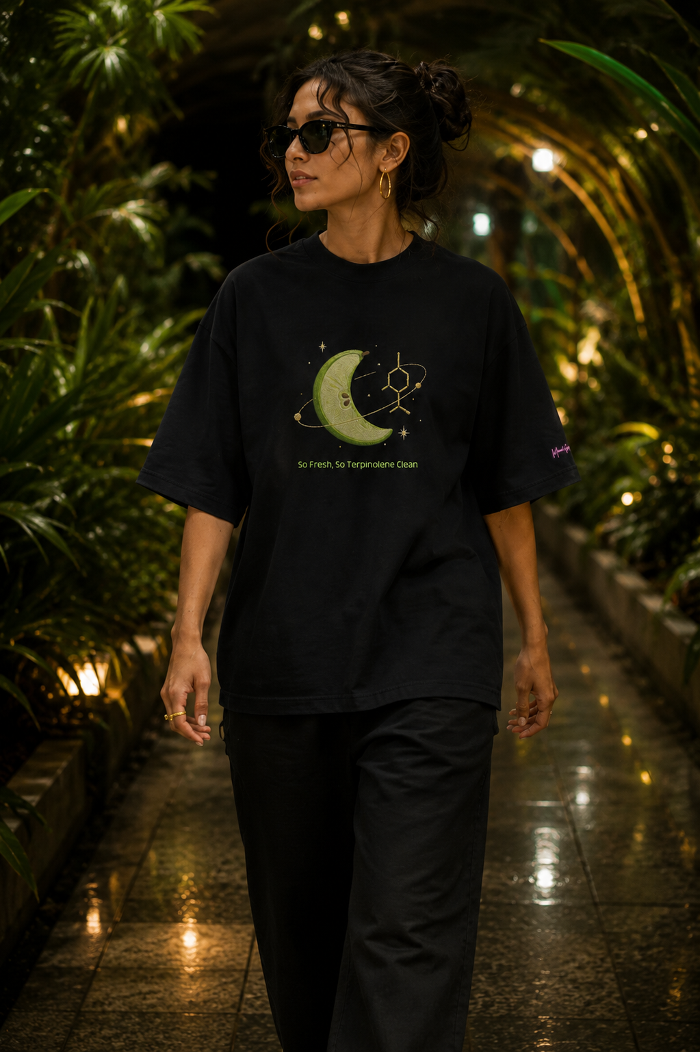 Woman wearing the black So Fresh, So Terpinolene Clean oversized tee while walking through a tropical conservatory.