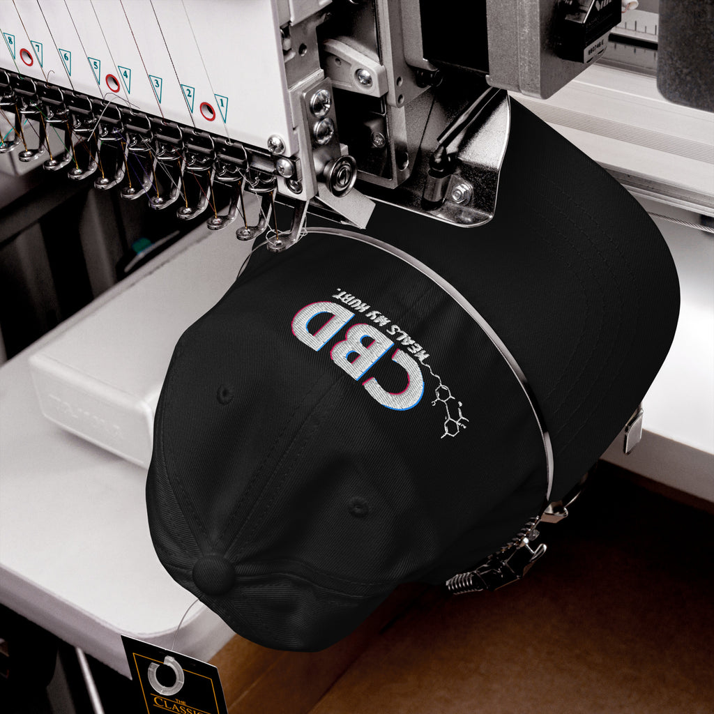 Black CBD Heals My Hurt dad hat secured inside a commercial embroidery machine as the front design is stitched.