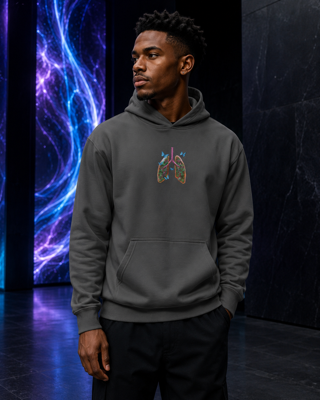 Male model wearing the charcoal Butterfly Lungs hoodie in a modern black gallery with a blue-violet cosmic light installation.