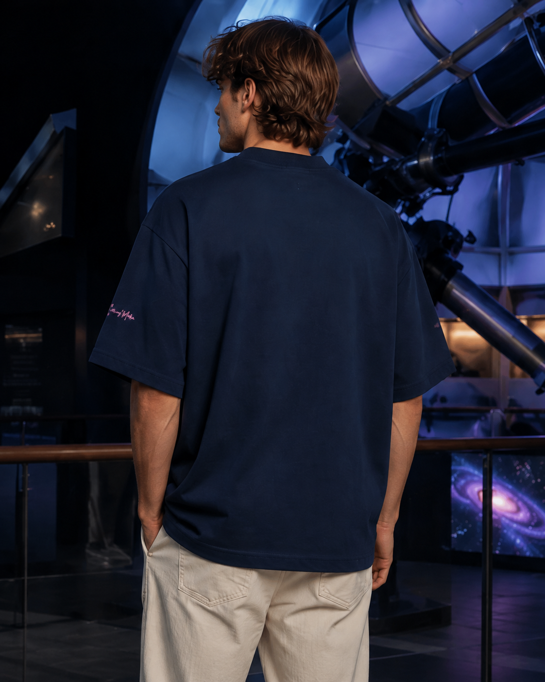 Back view of a man wearing the French navy oversized high-neck tee beside a large telescope in a blue-lit observatory.