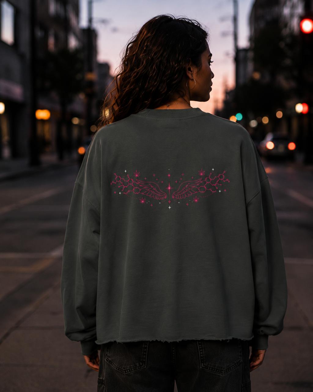 Back view of a woman wearing the Dark Gray Mandi Cruz Media raw edge boxy sweatshirt with pink celestial wing artwork on a city street at dusk.