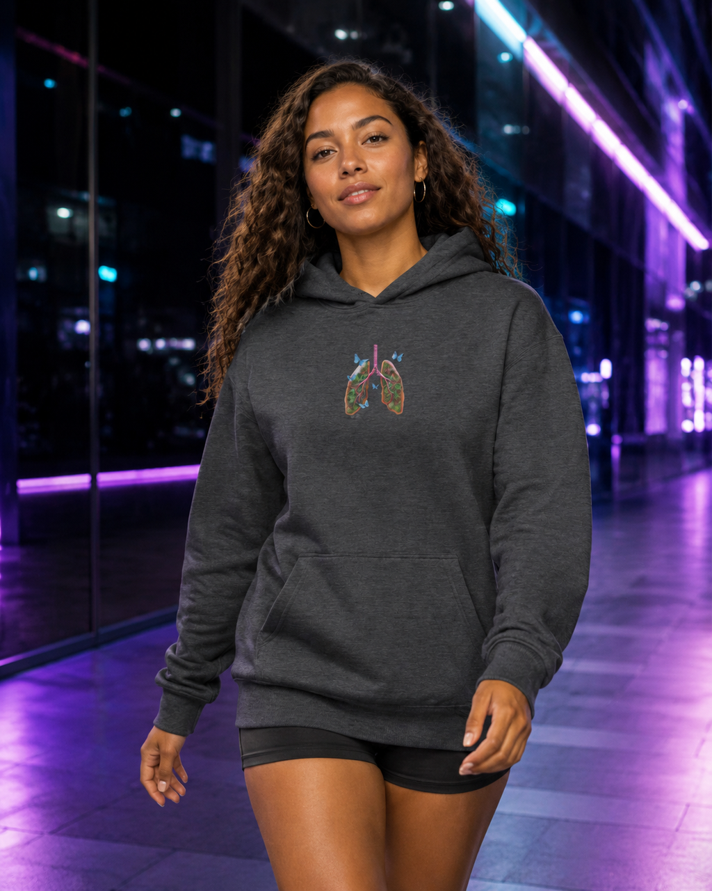 Young woman walking in a dark heather Butterfly Lungs hoodie through a sleek passage illuminated with violet and cyan lights.