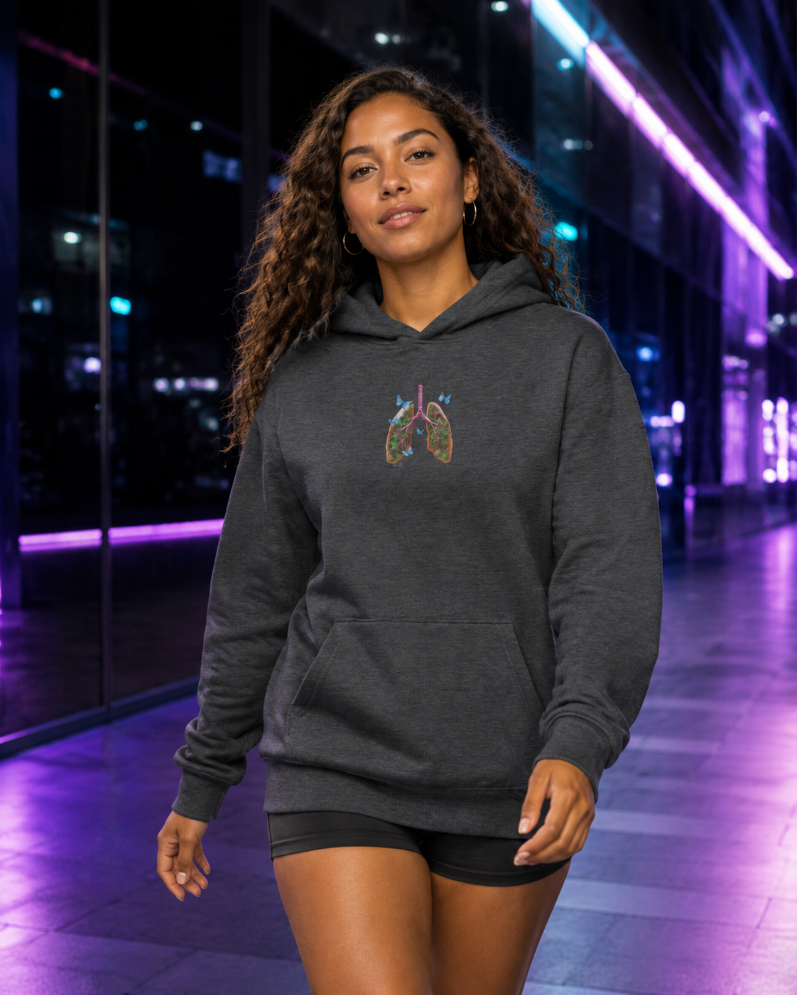 Young woman walking in a dark heather Butterfly Lungs hoodie through a sleek passage illuminated with violet and cyan lights.