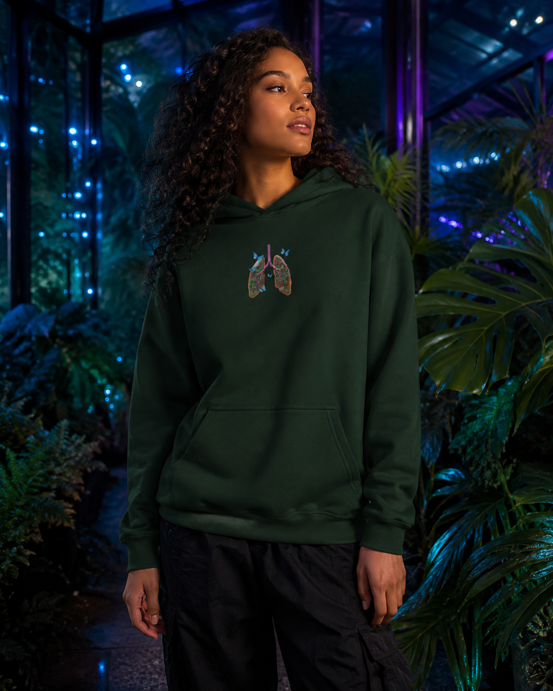 Young woman wearing the forest green Butterfly Lungs hoodie inside a bioluminescent botanical conservatory at night.