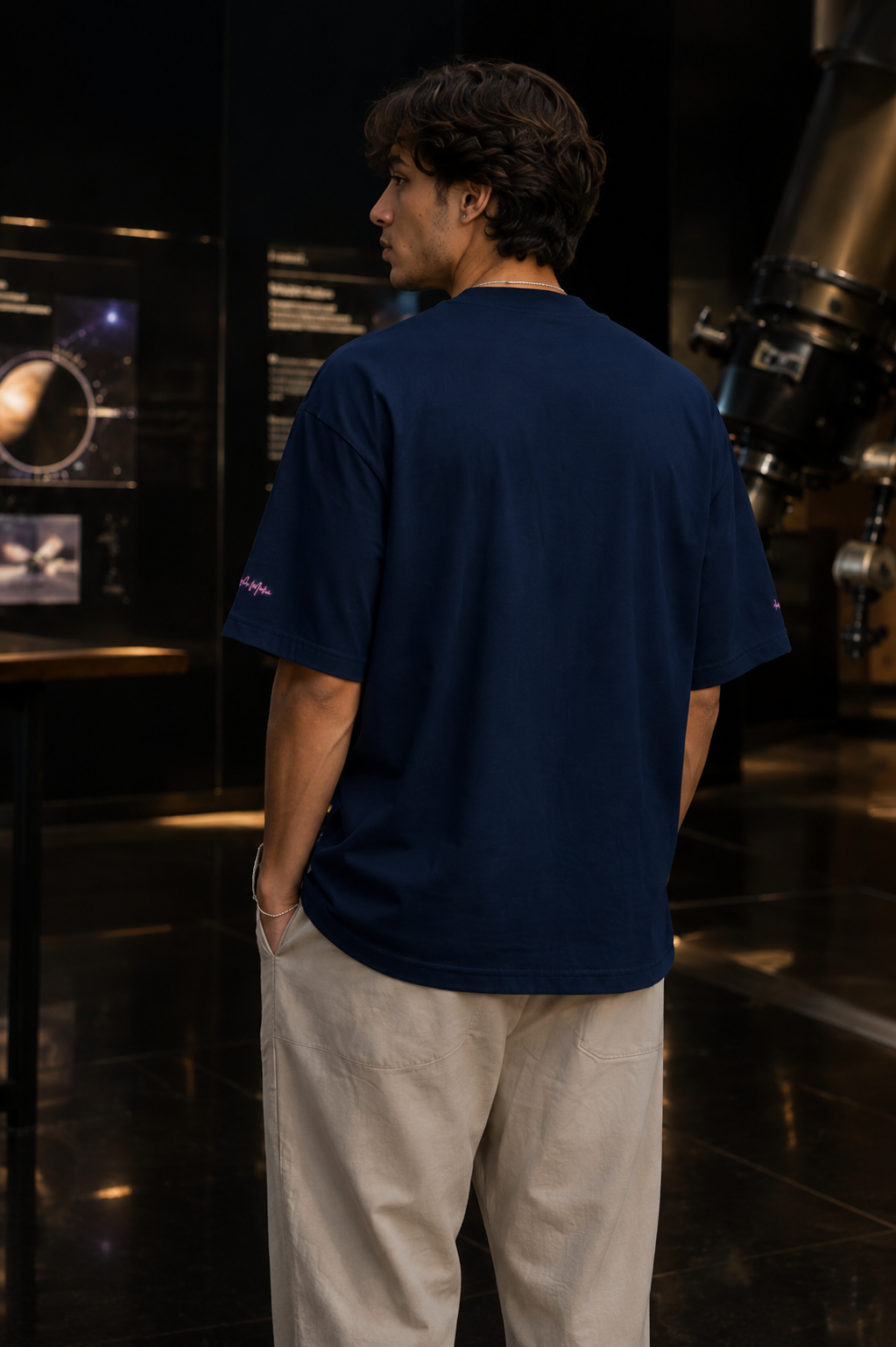 Back view of a man wearing the French-navy oversized organic tee inside a dark astronomy and science gallery