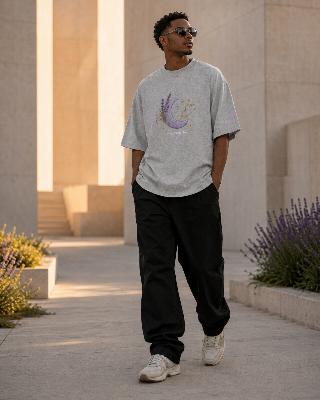 Man wearing the heather-grey In My Linalool Era oversized tee while walking through a modern courtyard with lavender planters.