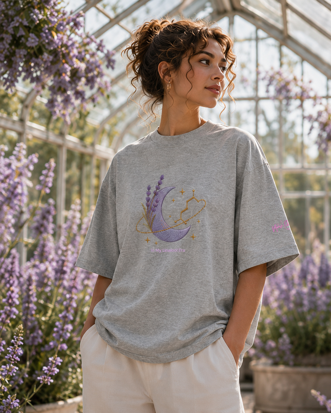 Woman wearing the heather-grey In My Linalool Era oversized tee in a sunlit greenhouse surrounded by lavender