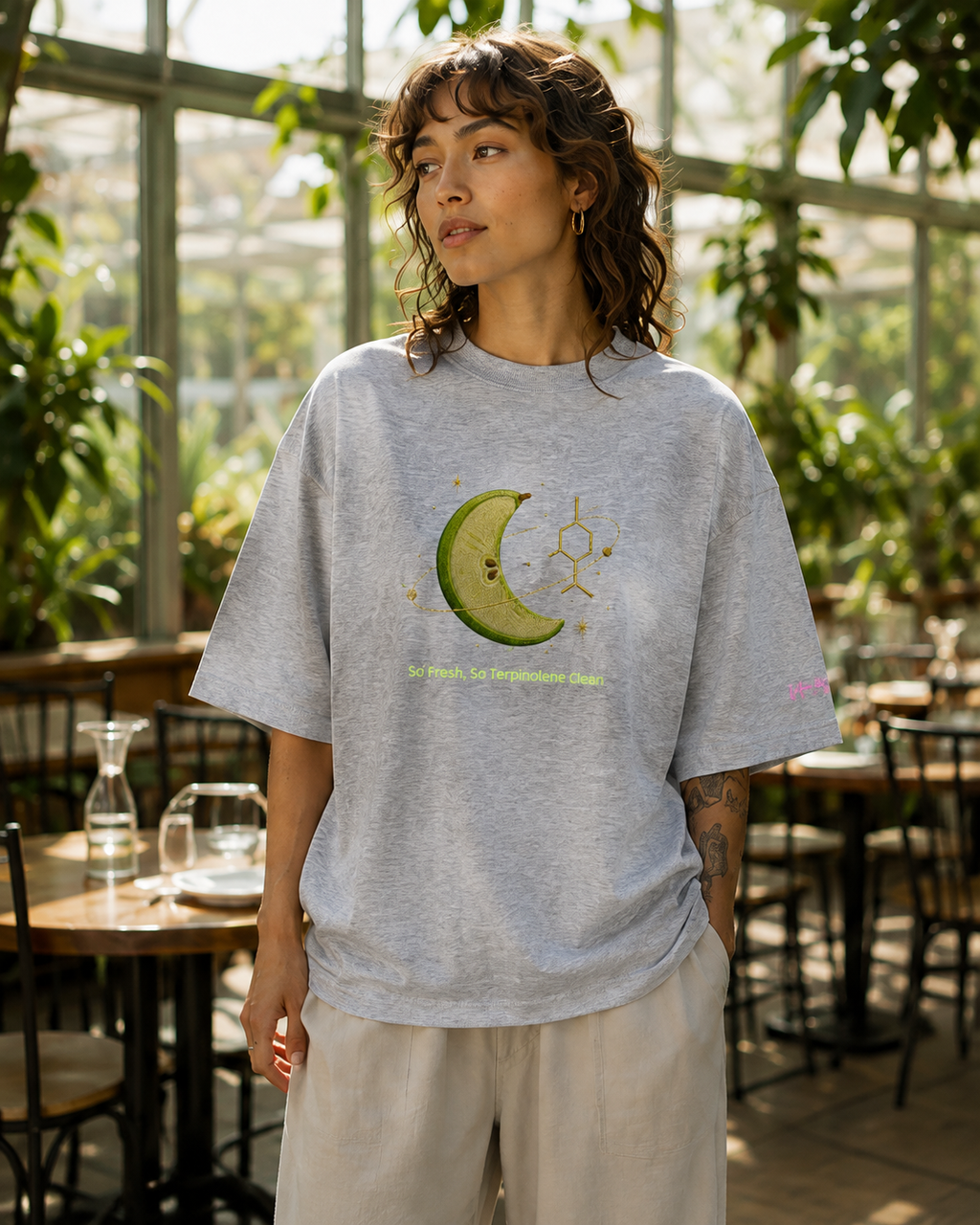 Woman wearing the heather-grey So Fresh, So Terpinolene Clean oversized organic tee in a sunlit botanical conservatory.