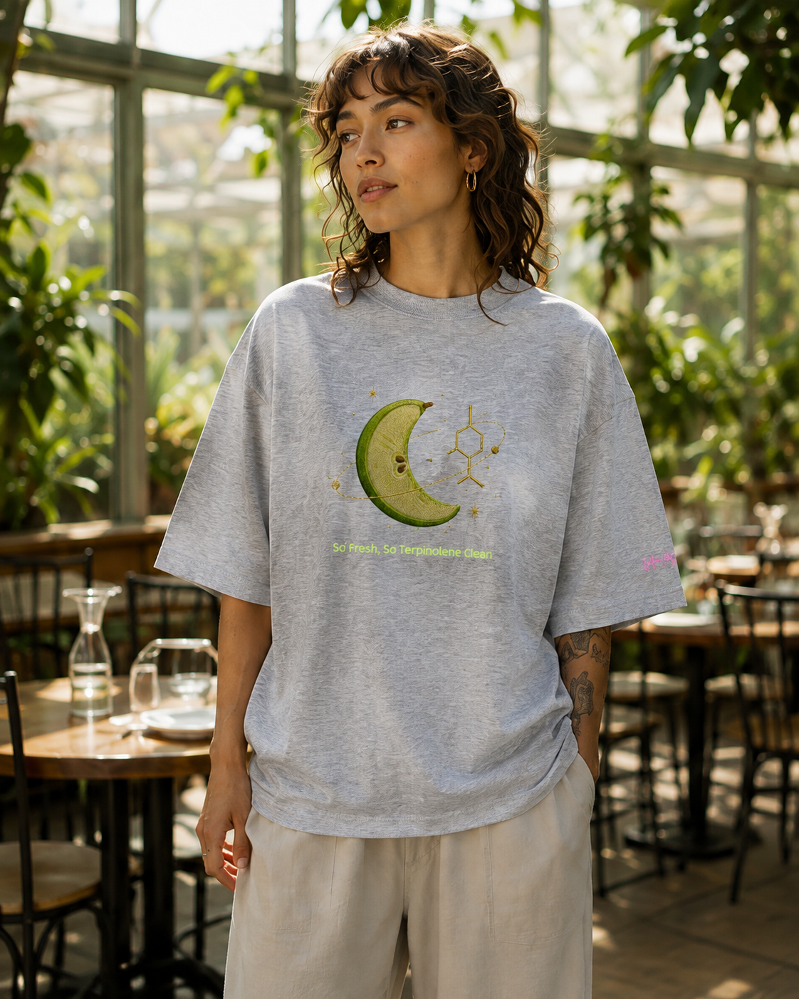 Woman wearing the heather-grey So Fresh, So Terpinolene Clean oversized organic tee in a sunlit botanical conservatory.