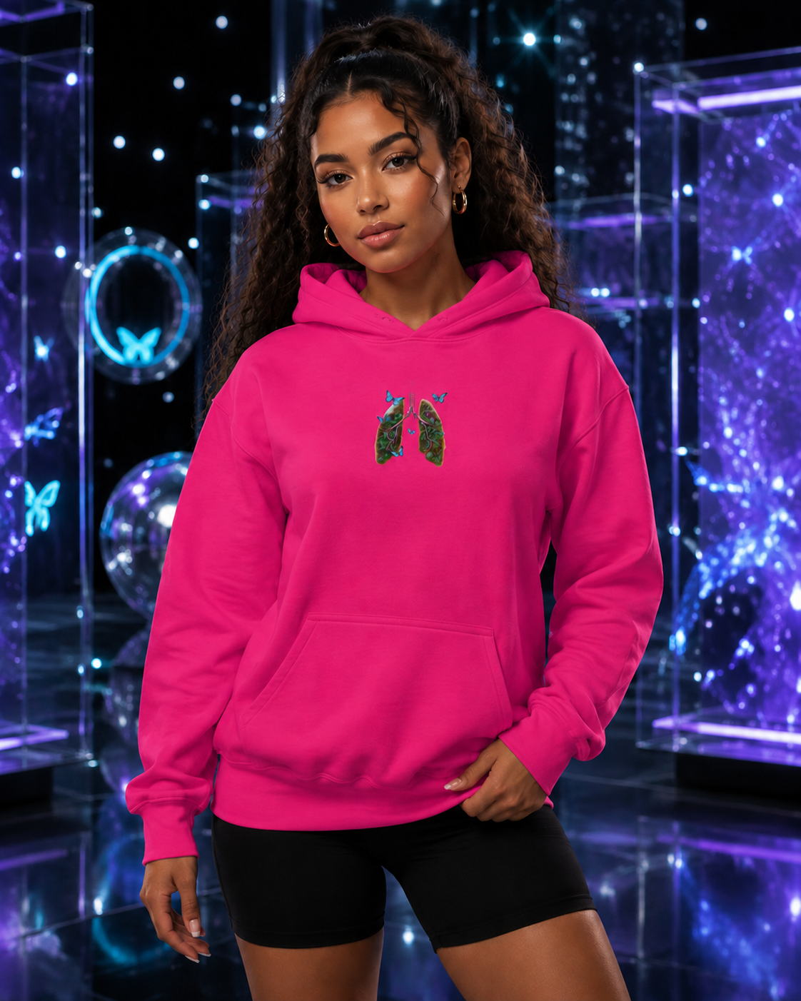 Young woman modeling the heliconia pink Butterfly Lungs hoodie with black shorts in a violet-and-cyan cosmic glass studio.