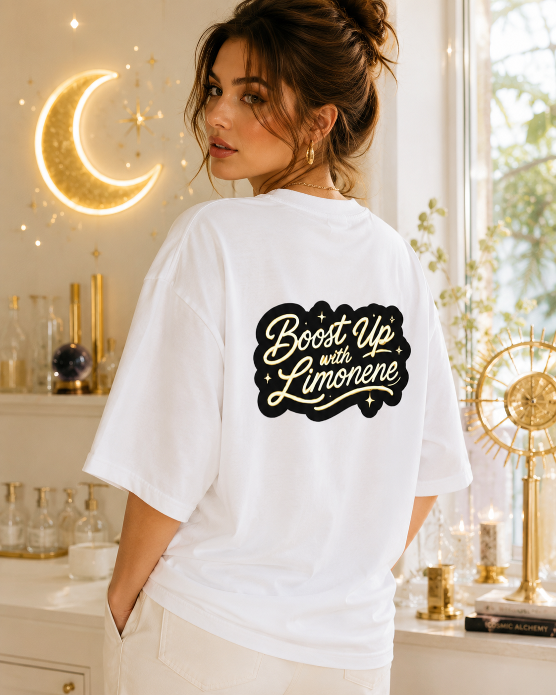 A woman wearing a white oversized t-shirt with a black graphic on the back that reads 'Boost Up with Limonene' in a cursive font, set in a warm, decorated room with a glowing moon lamp.Woman modeling the white oversized tee from the back, featuring the gold and black Boost Up with Limonene script design.