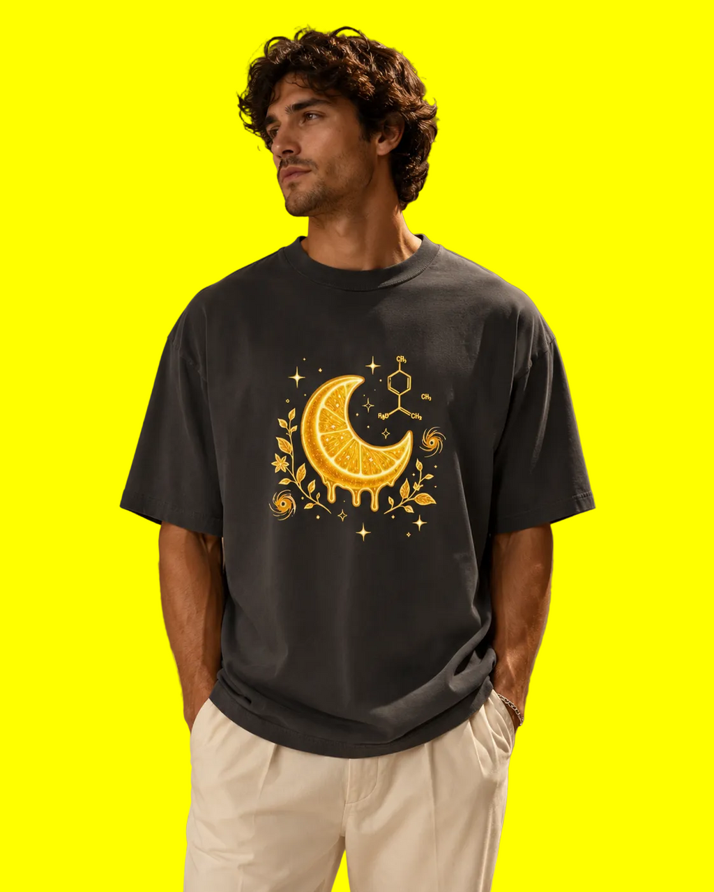 A man wearing a dark charcoal gray t-shirt with a yellow and orange citrus moon graphic, paired with off-white trousers, standing in a warm, modern architectural setting.