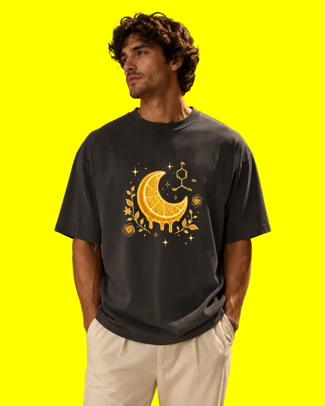 A man wearing a dark charcoal gray t-shirt with a yellow and orange citrus moon graphic, paired with off-white trousers, standing in a warm, modern architectural setting.