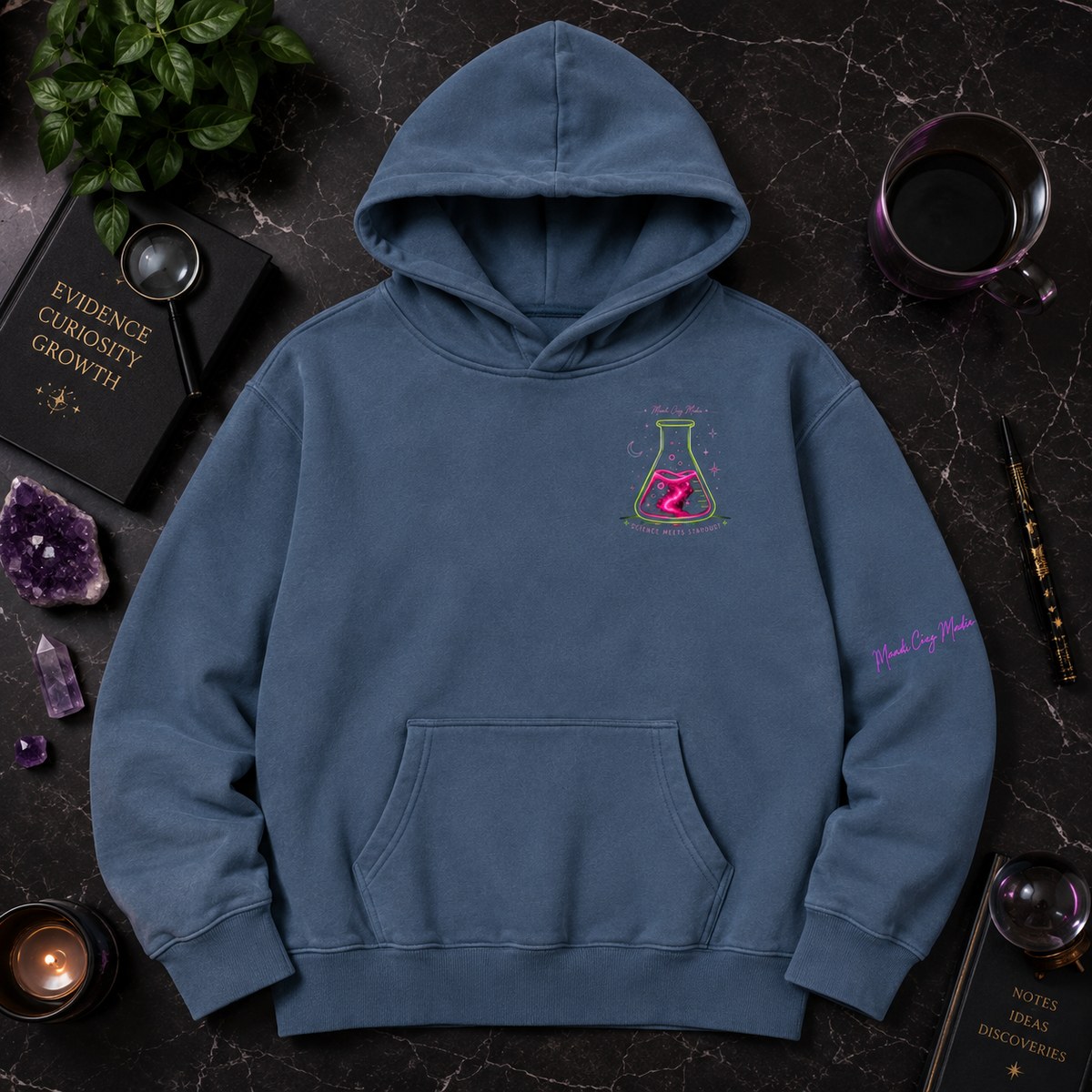 Blue Mandi Cruz Media hoodie shown from the front with neon flask chest graphic and sleeve detail, styled with crystals, greenery, journal and dark celestial accessories.