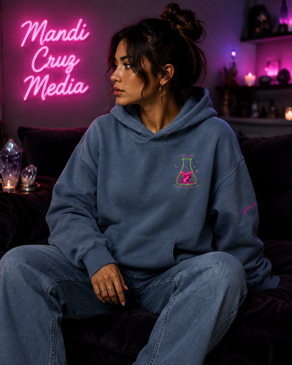 Woman modeling the blue Mandi Cruz Media oversized hoodie with small neon flask chest graphic and signature sleeve detail in a softly lit pink and purple studio.
