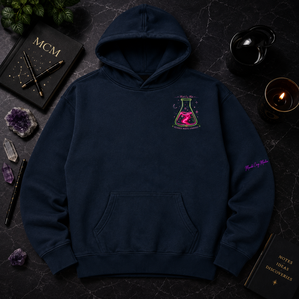 Navy Mandi Cruz Media oversized hoodie displayed in a premium dark flat lay with neon flask chest artwork, crystals, candle, journal and celestial accents.