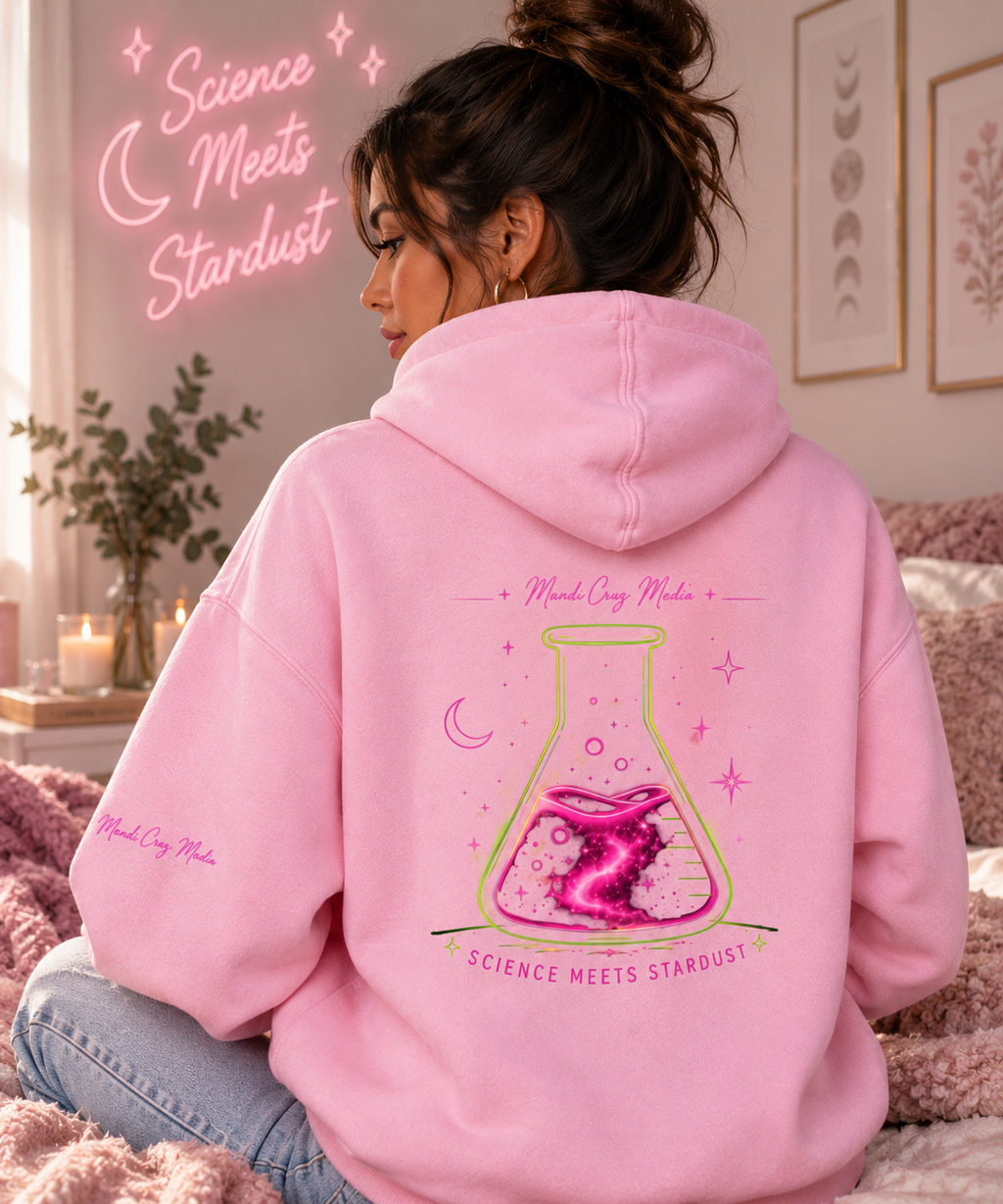 Woman wearing a pink Mandi Cruz Media oversized hoodie from the back in a cozy interior, displaying the large Science Meets Stardust flask graphic and sleeve branding.