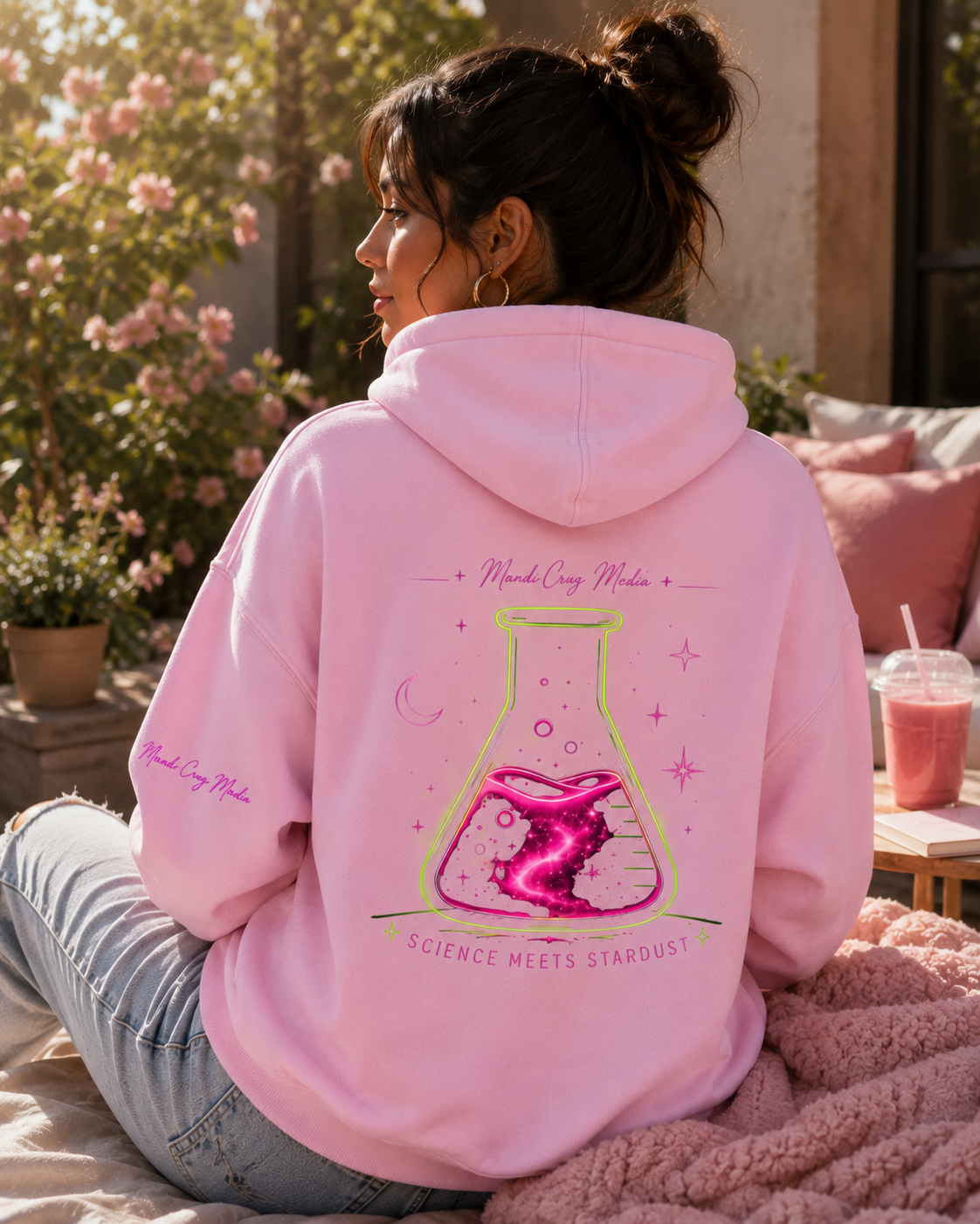 Woman wearing a pink Mandi Cruz Media hoodie from the back in a sunlit outdoor setting, showcasing the large neon flask, celestial details, and Science Meets Stardust design.