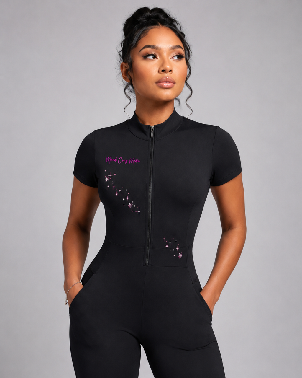 Close-up of the black quarter-zip jumpsuit showing its short sleeves, fitted waist, front pockets, pink Mandi Cruz Media logo, and constellation design.