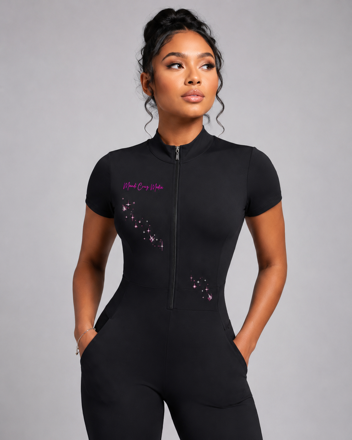 Close-up of the black quarter-zip jumpsuit showing its short sleeves, fitted waist, front pockets, pink Mandi Cruz Media logo, and constellation design.