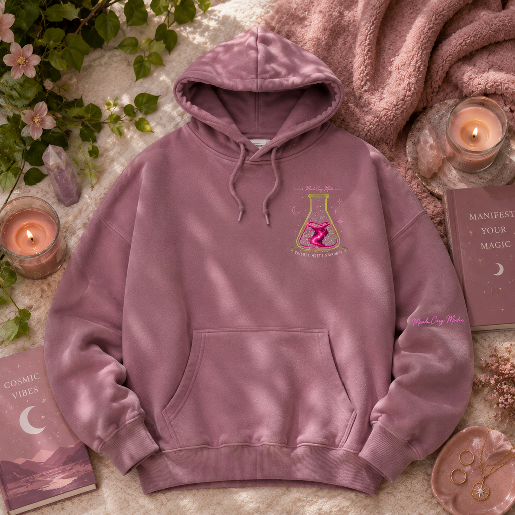 Dusty mauve Mandi Cruz Media oversized hoodie shown in a botanical flat lay with a small Science Meets Stardust flask chest graphic, sleeve branding, crystals, candles, and greenery.