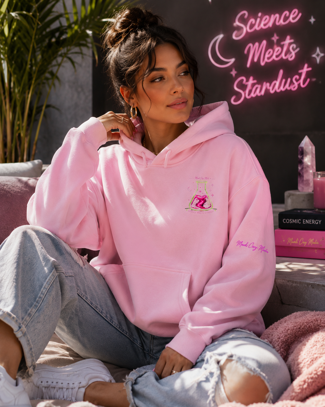 Woman wearing a pink Mandi Cruz Media oversized hoodie featuring a small Science Meets Stardust flask graphic on the front chest and pink sleeve branding.