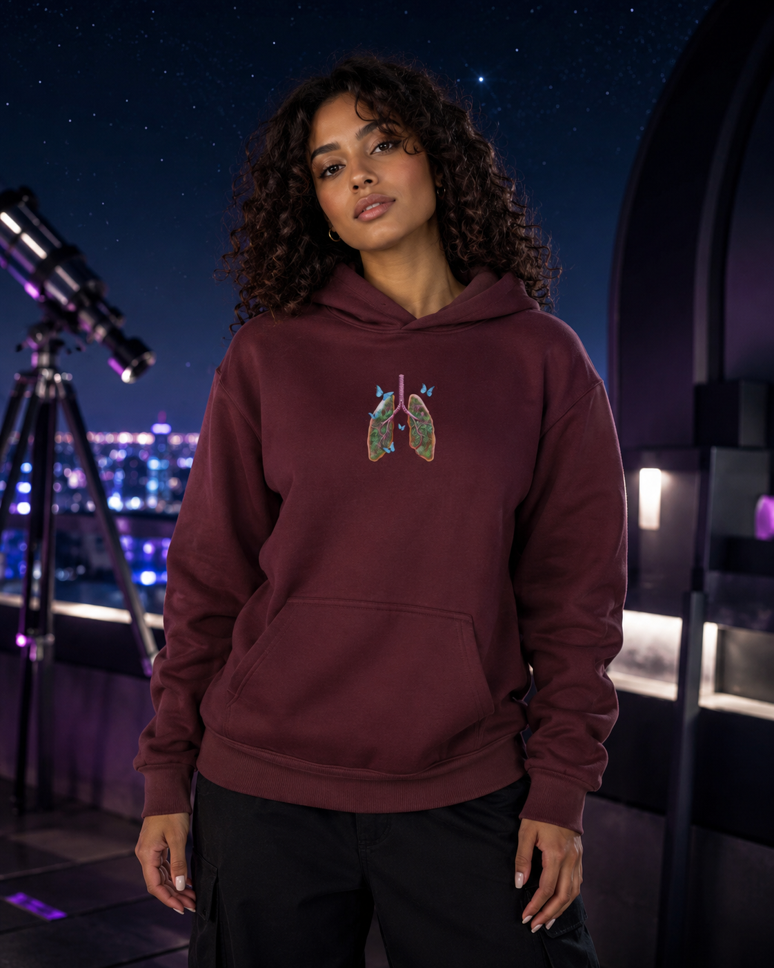 Young woman modeling the maroon Butterfly Lungs hoodie on a rooftop observatory with a telescope, stars and city lights.