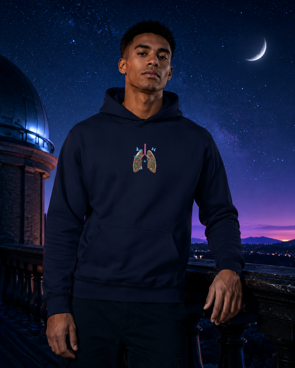 Male model wearing the navy Butterfly Lungs hoodie on a nighttime observatory terrace beneath stars and a crescent moon.