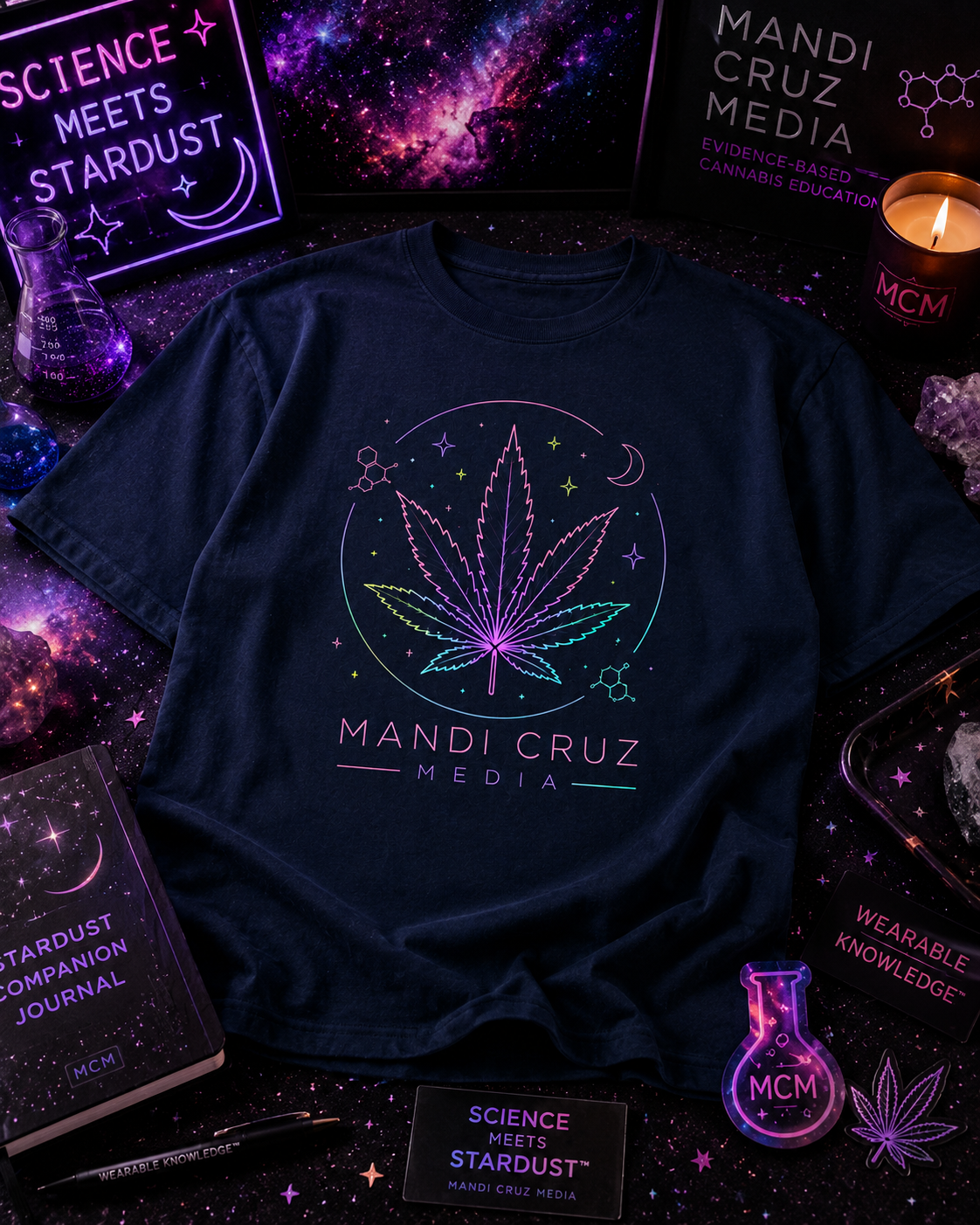 A navy blue graphic t-shirt featuring a glowing neon-style cannabis leaf design with the text 'MANDI CRUZ MEDIA' underneath, styled in a dark cosmic flat-lay with books, crystals, and themed props.