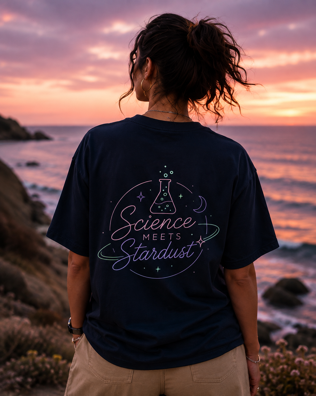 A woman stands on a rocky beach at sunset facing the ocean, wearing a navy blue t-shirt with a colorful graphic on the back that reads 'Science Meets Stardust'.
