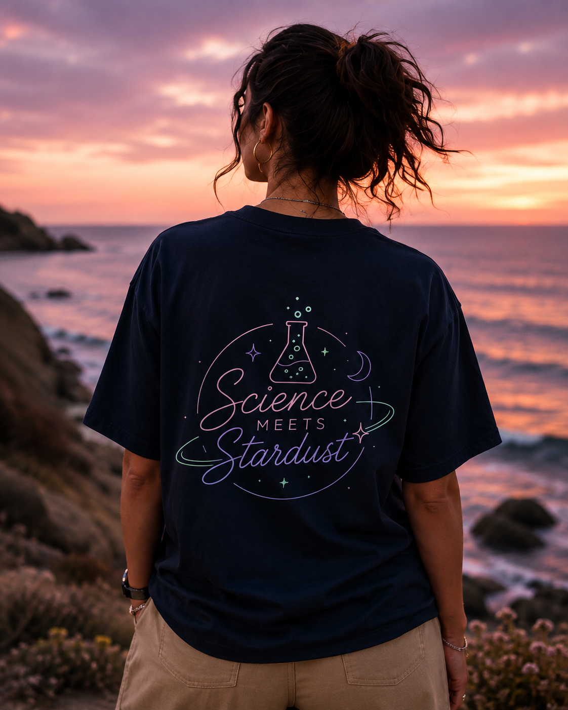A woman stands on a rocky beach at sunset facing the ocean, wearing a navy blue t-shirt with a colorful graphic on the back that reads 'Science Meets Stardust'.