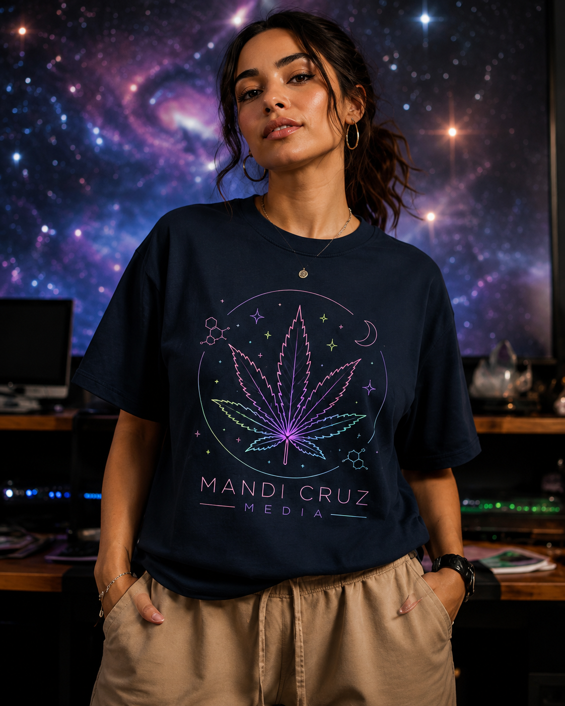 A woman wearing a navy blue graphic t-shirt with a purple and pink cannabis leaf design and the text 'MANDI CRUZ MEDIA', paired with khaki pants, set against a dark room with a starry galaxy screen background.