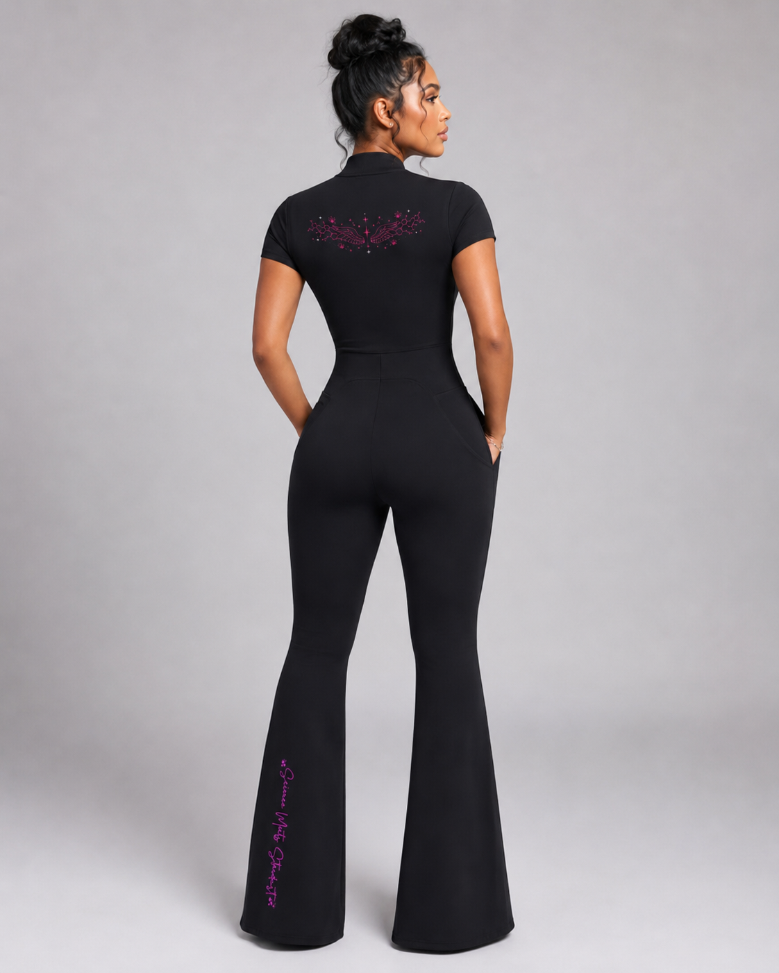 Model wearing the black fitted flared-leg jumpsuit from the back, displaying pink molecular wings and vertical Science Meets Stardust script.