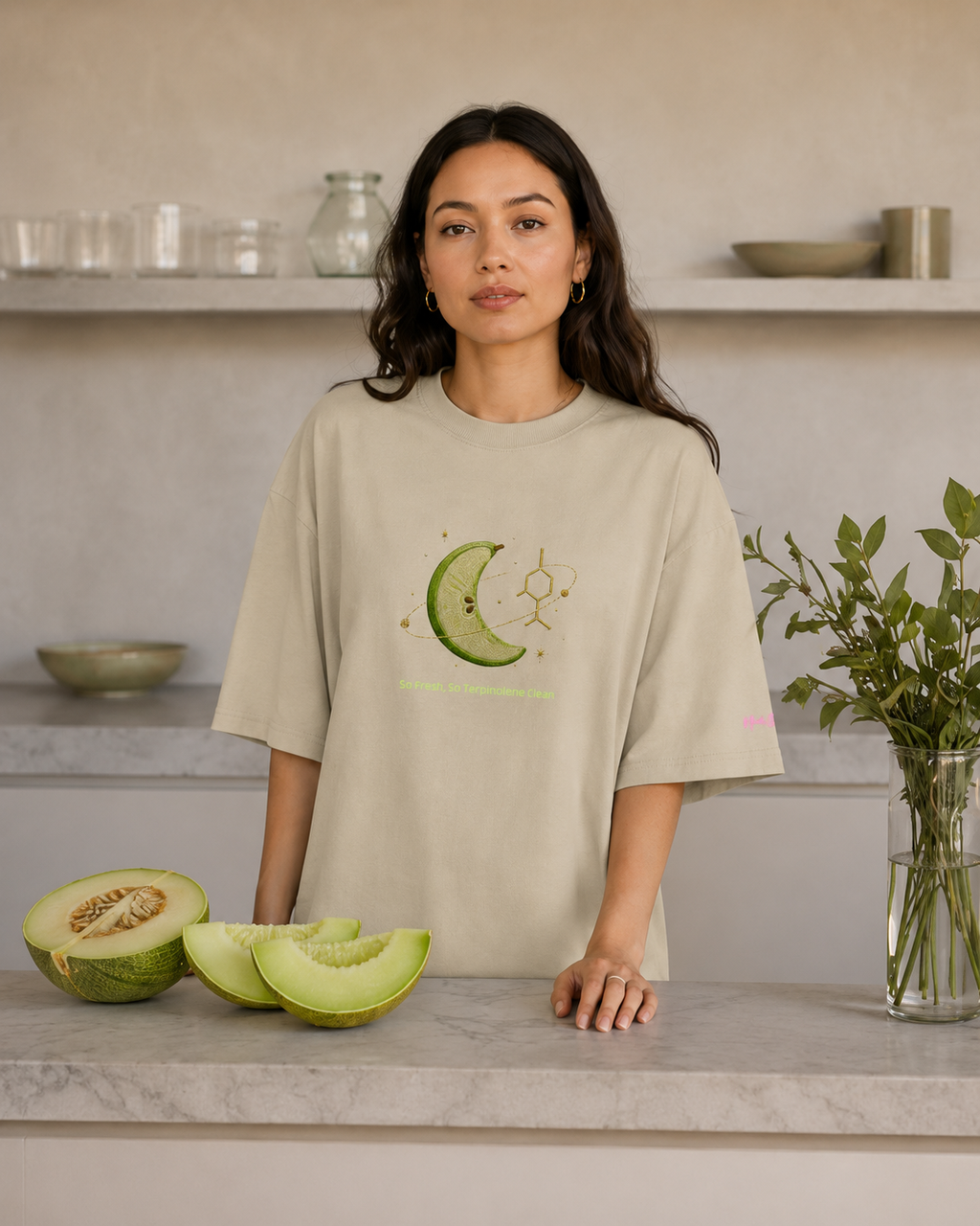 Woman wearing the stone So Fresh, So Terpinolene Clean oversized tee beside fresh melon in a modern kitchen.