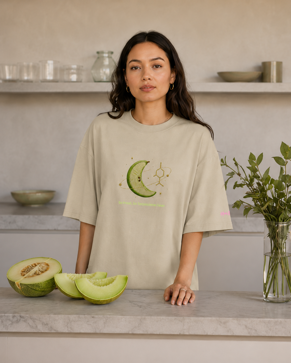 Woman wearing the stone So Fresh, So Terpinolene Clean oversized tee beside fresh melon in a modern kitchen.