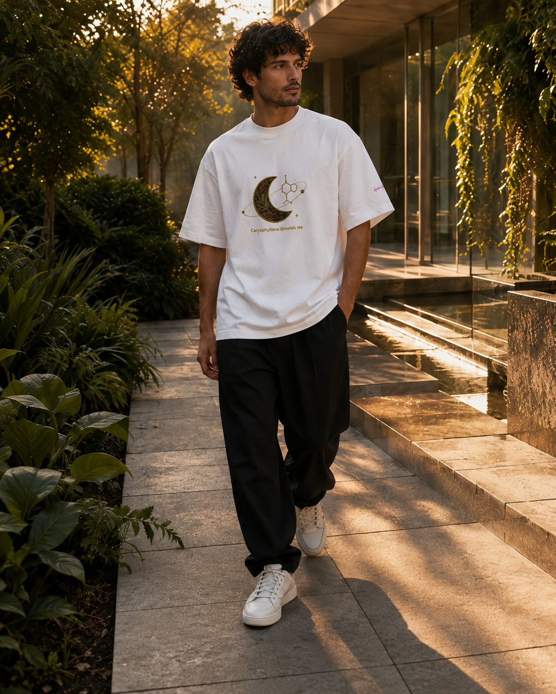 Man wearing the white Caryophyllene Grounds Me oversized tee while walking through a modern garden at golden hour.