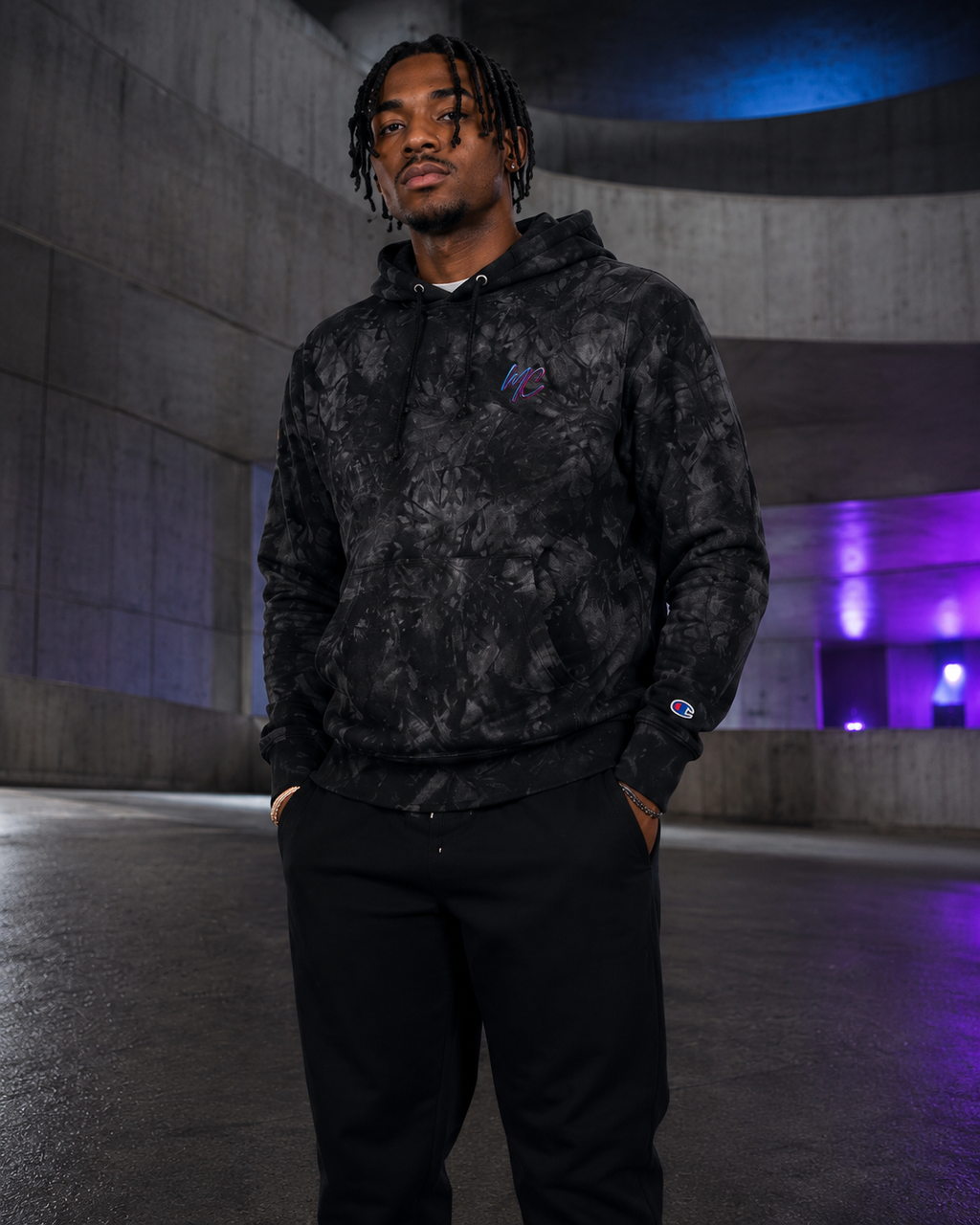 Male model wearing the black-and-charcoal Champion tie-dye hoodie in a modern concrete architectural setting.