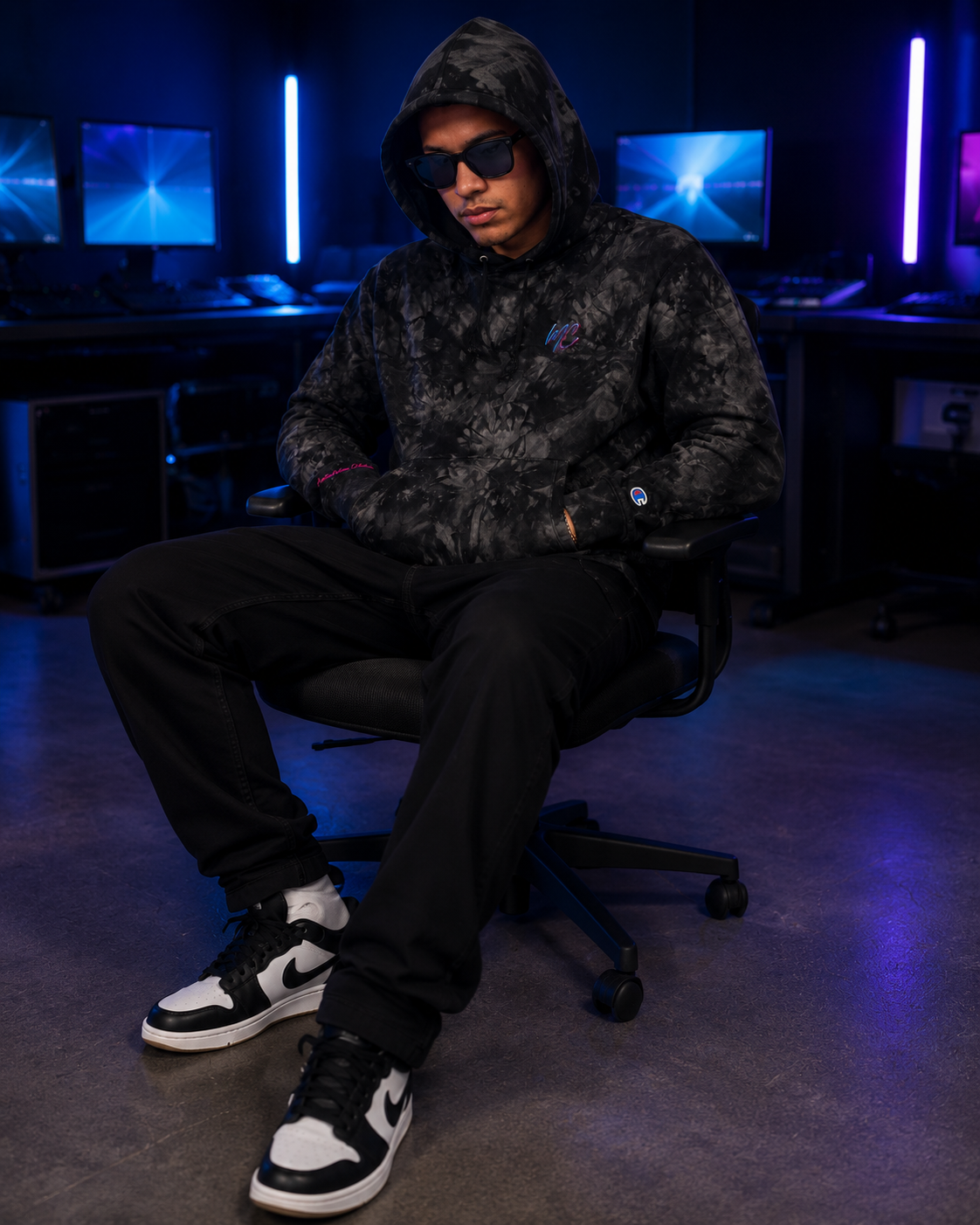 Seated male model wearing the Champion tie-dye hoodie with the hood raised in a cyan-and-violet creative studio.