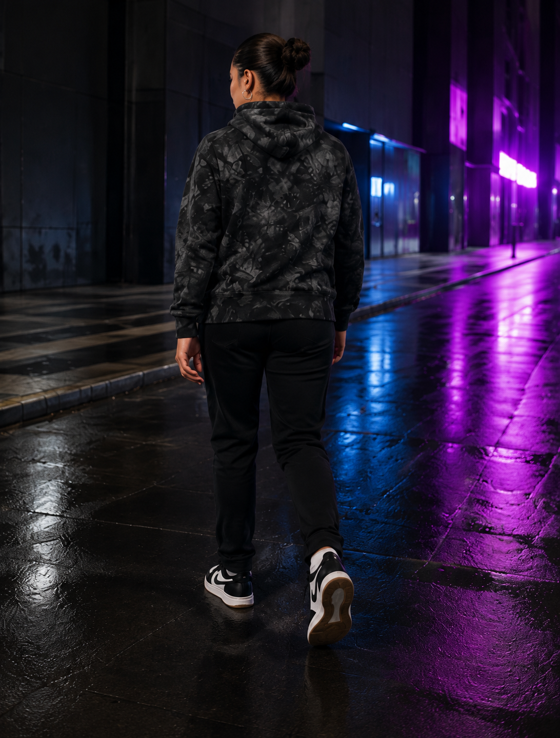Female model showing the blank back of the Champion tie-dye hoodie while walking along a wet city street at night.
