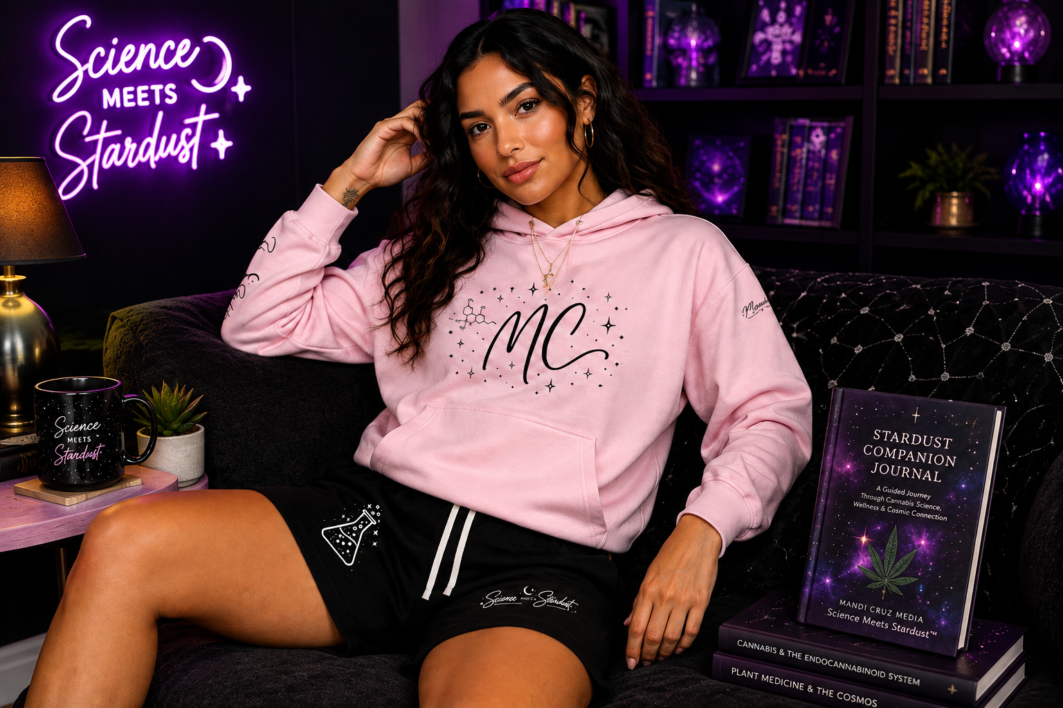 Woman wearing a pink Mandi Cruz Media hoodie and black Science Meets Stardust™ shorts beside the Stardust Companion Journal in a purple cosmic lounge.