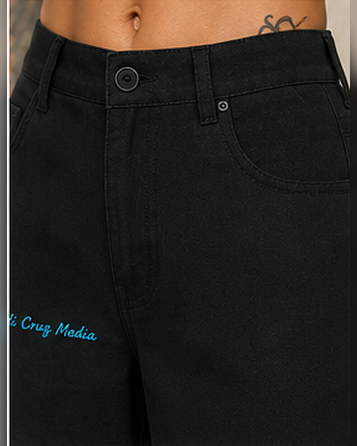 Close-up of a person wearing black denim shorts with a small blue embroidered look logo on the left leg, showing part of the waist and hip.