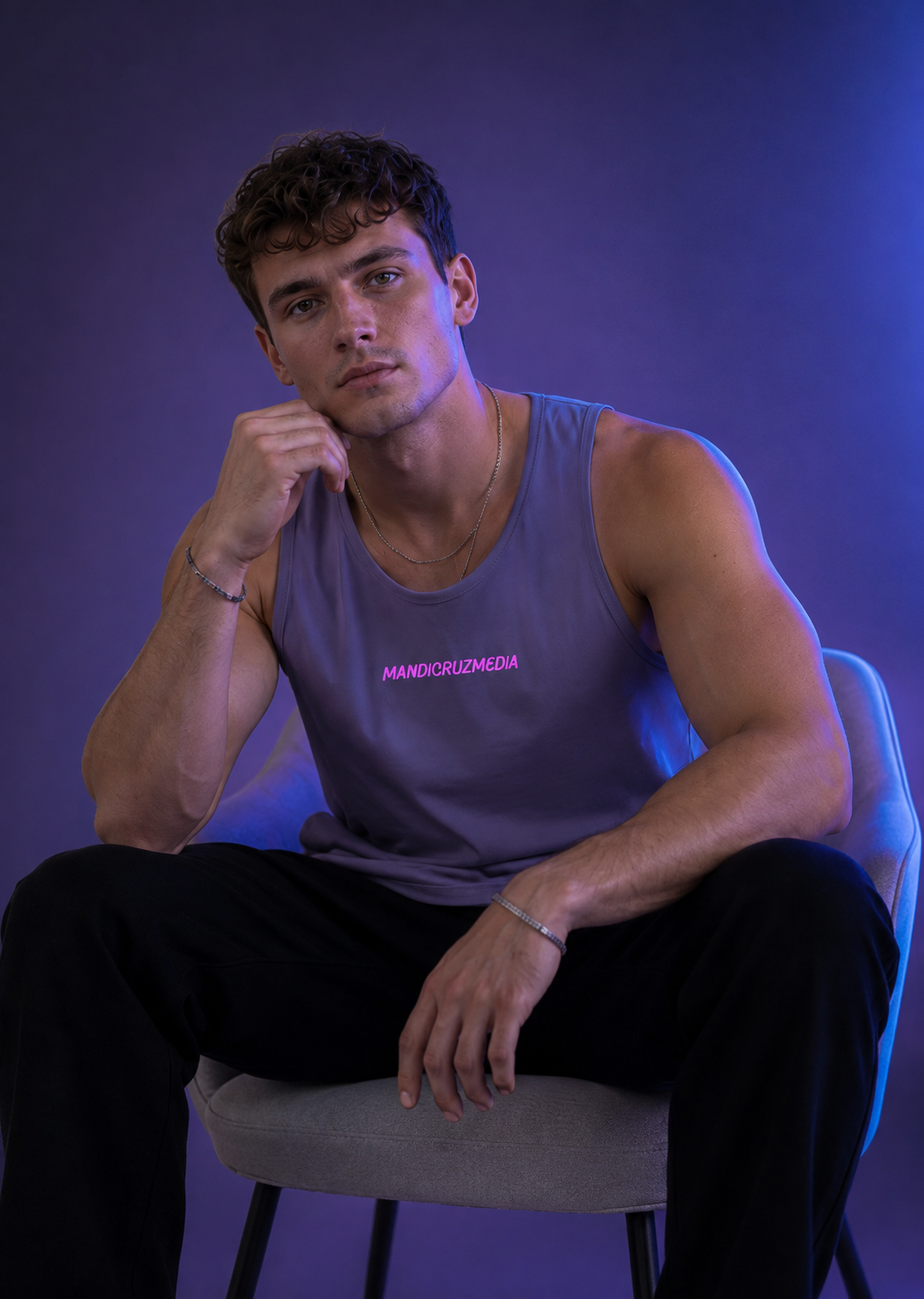 Seated male model wearing the Harbor Blue Mandi Cruz Media premium tank under violet and blue studio lighting.